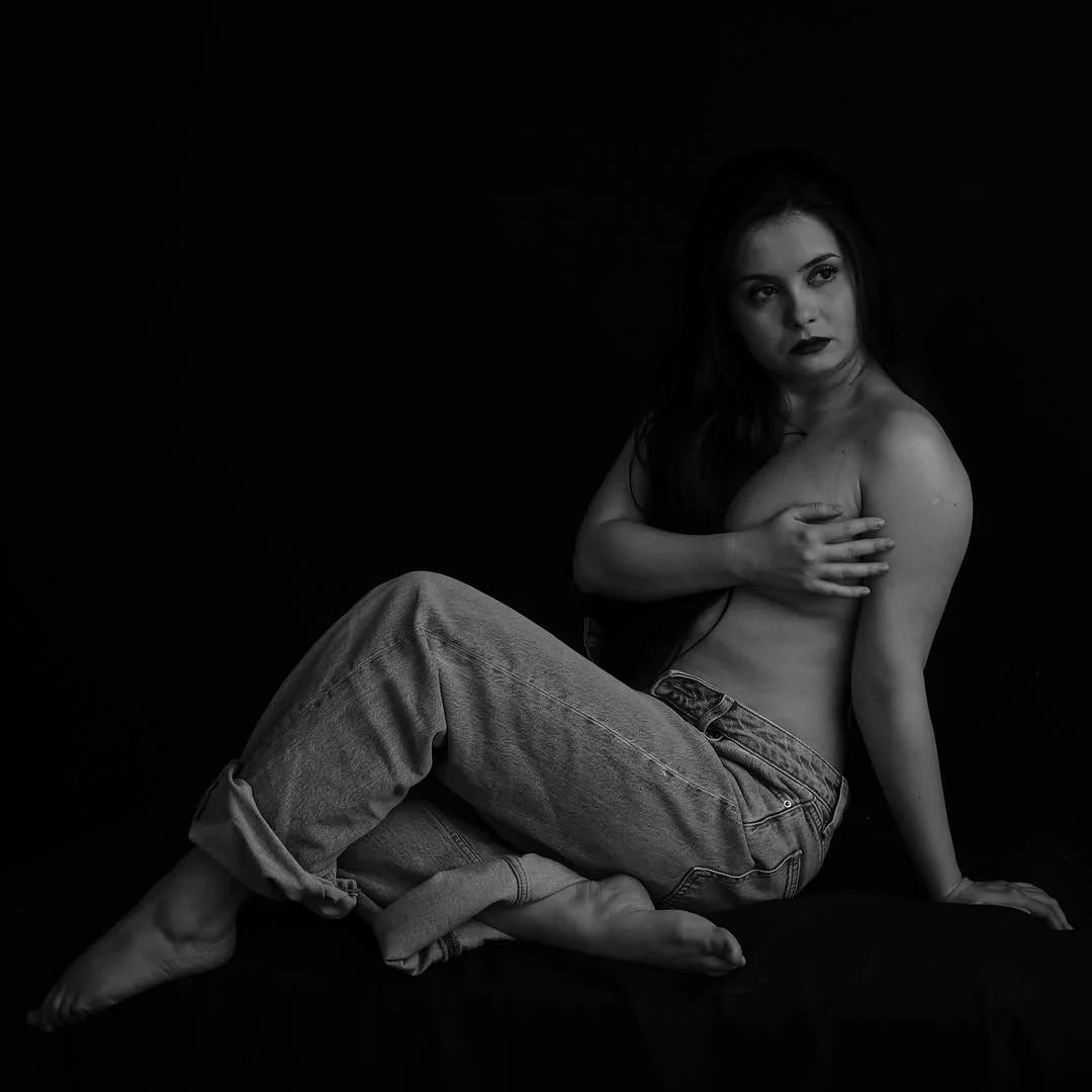 A woman in jeans sits turned toward the camera in black and white, lit from the side against a black backdrop