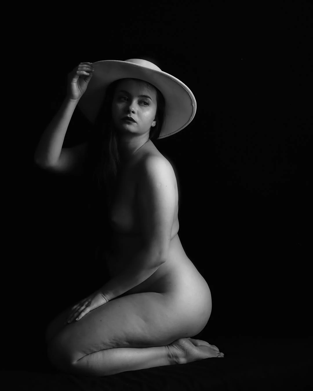 A woman in a wide-brimmed hat kneels in black and white, one hand on the brim, lit softly from one side