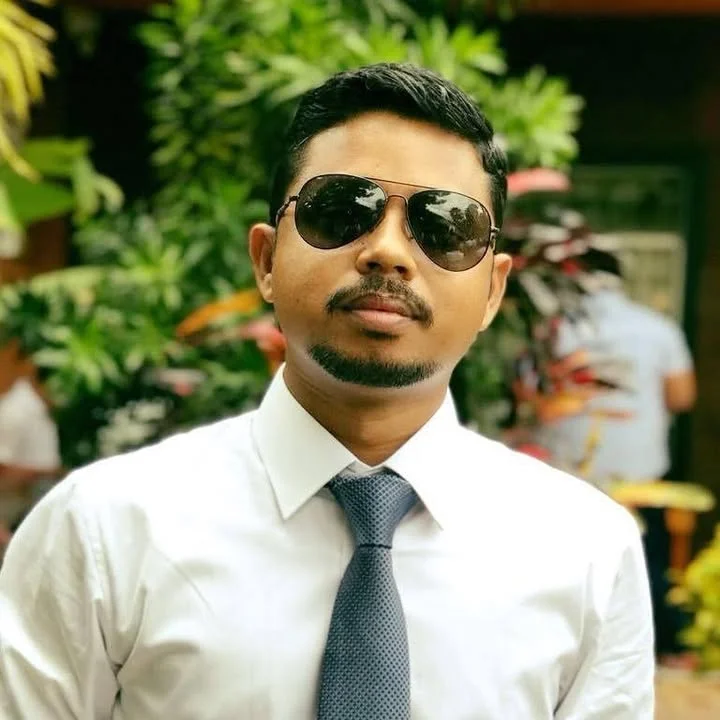 Hirak Choudhury wearing sunglasses, a white shirt and a grey tie, with green foliage behind him
